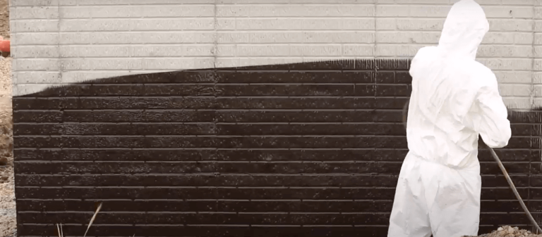 A person in a white protective suit and hood paints a brick wall with black paint using a long-handled roller. The upper part of the wall remains white, while the lower part is freshly coated black.