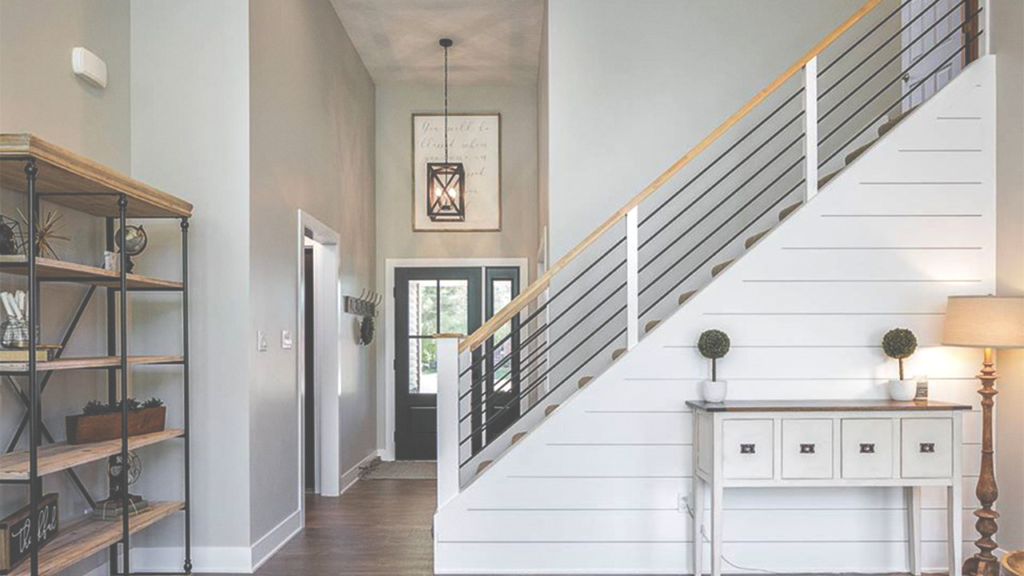 A modern entryway in a KRM Custom Homes design features wooden flooring and a white staircase with metal railings leading to the upper floor. On the right, there's a console table with a lamp and decorative items. To the left, a black metal and wood shelving unit stands out. The front door is visible in the background.