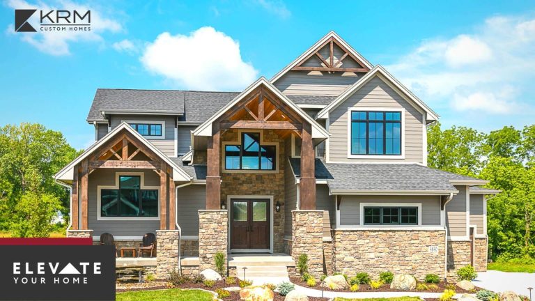 A modern, two-story house with grey siding, large windows, and wooden beams framing the entrance. The home features a neatly landscaped front yard with shrubs and stone accents. The logo for KRM Custom Homes and the text "Elevate Your Home" are positioned in the lower left corner.