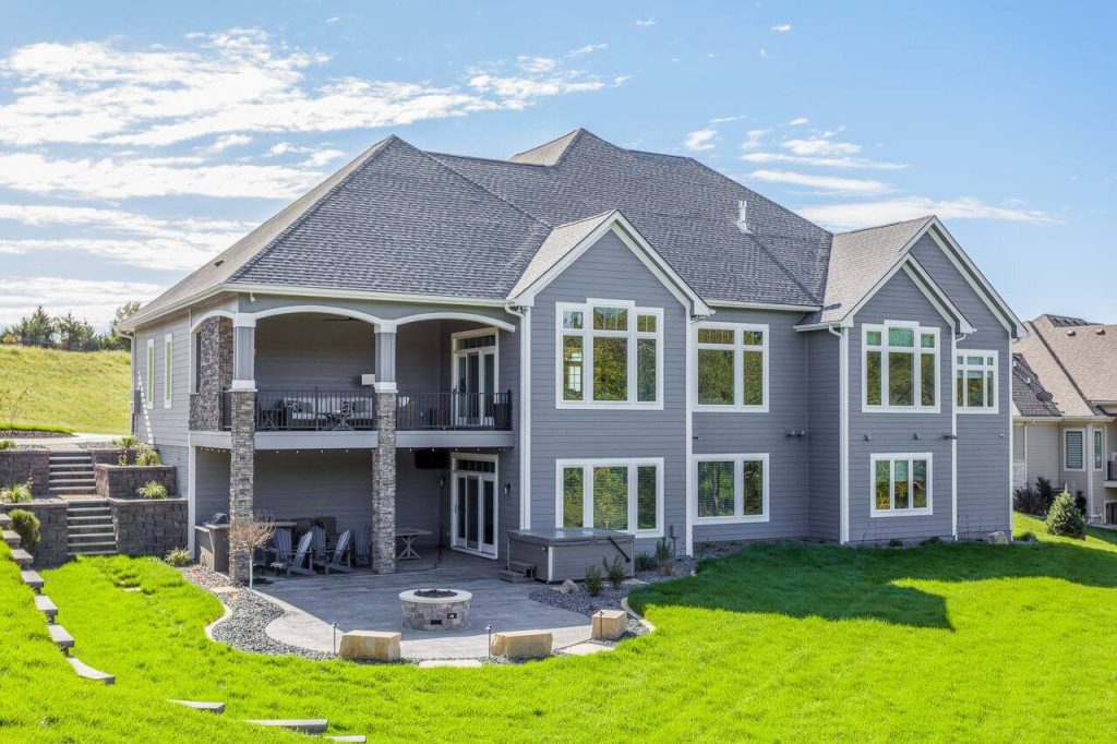Large modern gray two-story house with multiple windows, two balconies, and a stone fire pit in the backyard.