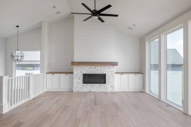 A modern, white living room with a vaulted ceiling, featuring a central fireplace with a stone surround. The fireplace is flanked by built-in cabinets. A large window fills the right wall, and a ceiling fan with dark blades hangs in the center of the room.