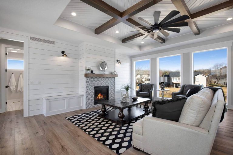 A modern living room features white shiplap walls, a large black ceiling fan, and a fireplace with patterned tile surround. There's a beige sofa, black chairs, and a rug with a geometric pattern. Large windows offer a clear view of the outdoors.