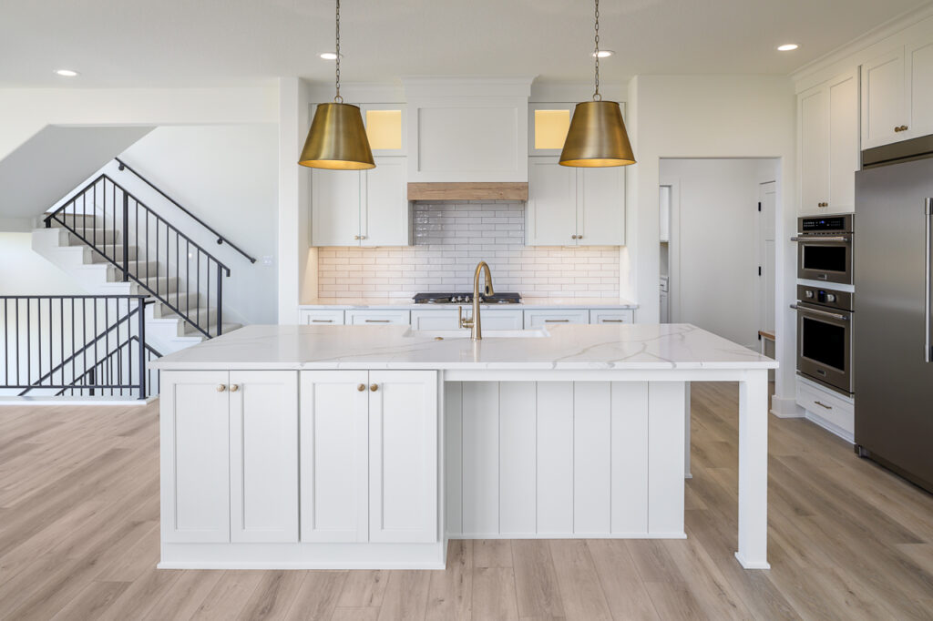 A modern kitchen with white cabinetry, a large white central island, and a marble countertop. Two gold pendant lights hang above the island. The backsplash features white subway tiles. Stainless steel appliances, including a refrigerator and double oven, are visible.