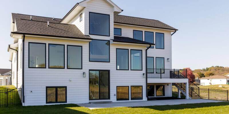 A modern, multi-story house with white siding and large windows. The house features a dark roof and a spacious backyard with a manicured lawn. A staircase leads from an elevated deck to the lawn. The sky is clear, and neighboring houses are visible in the distance.