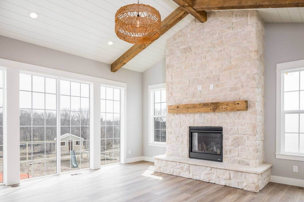 Bright living room with large windows, stone fireplace, wooden mantel, and modern hanging light fixture.