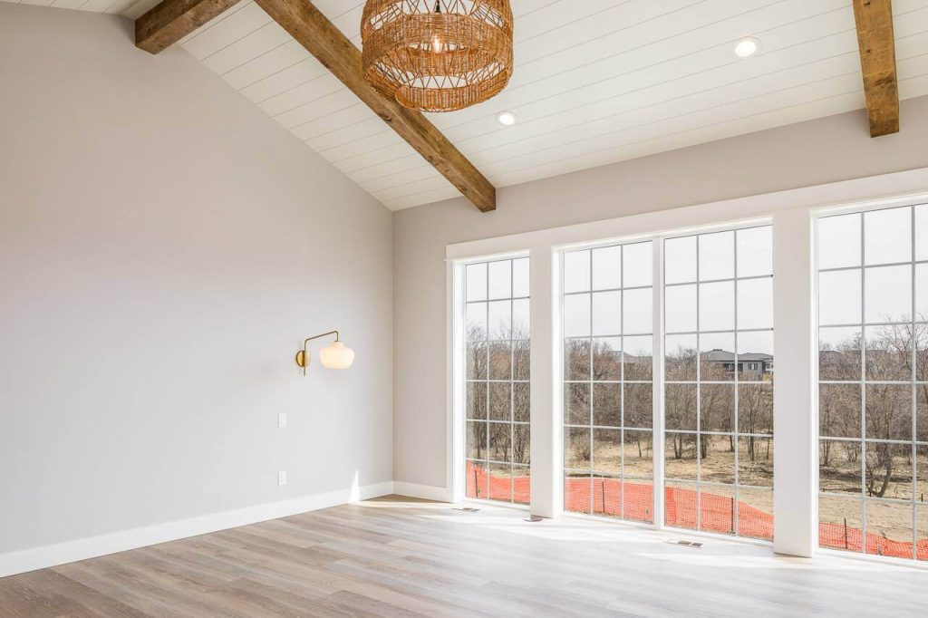 Bright empty room with wood beams, large windows, and a modern light fixture, overlooking a wooded view.
