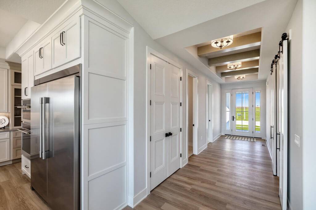 A modern hallway with light wood flooring and white walls, featuring recessed ceiling lights and exposed beams. A large stainless steel refrigerator is visible on the left, adjacent to a kitchen area. The hallway leads to a glass door with a view of greenery outside.