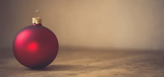 A single red Christmas ornament with a gold cap and hook sits on a wooden surface. The background is softly blurred, creating a warm, festive atmosphere.