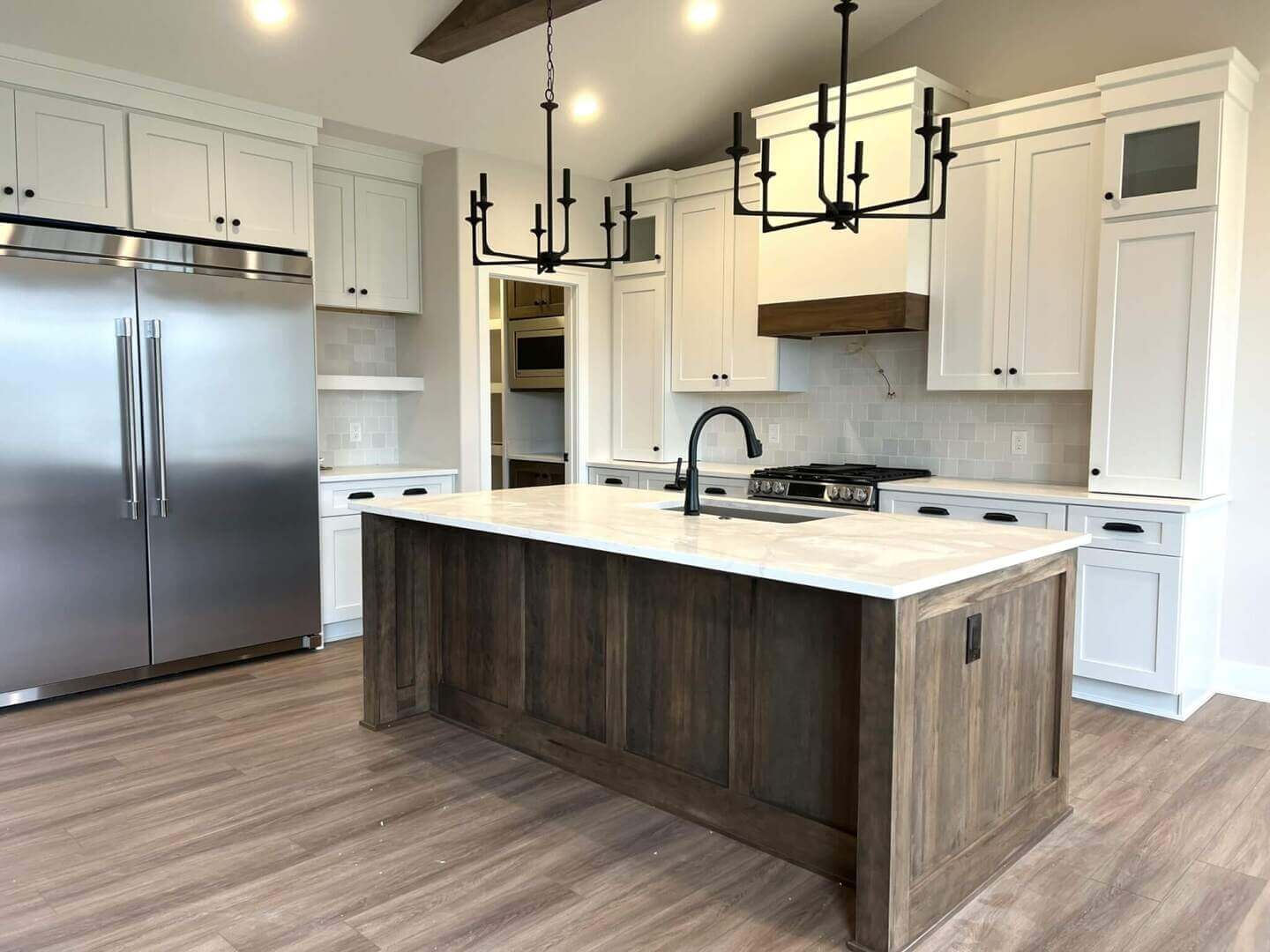 A modern kitchen with white cabinets, a large stainless steel refrigerator, and a central island with a wood finish and white countertop. Black chandeliers hang from the ceiling. The floor is wooden, and the walls are a light color.