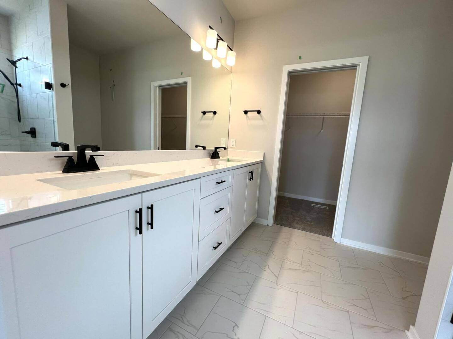 A modern bathroom with white cabinets, dual sinks, black faucets, and a large wall mirror with overhead lighting. The floor is tiled in white, and a door leads to a walk-in closet. Another door to the side appears to lead to a shower area.