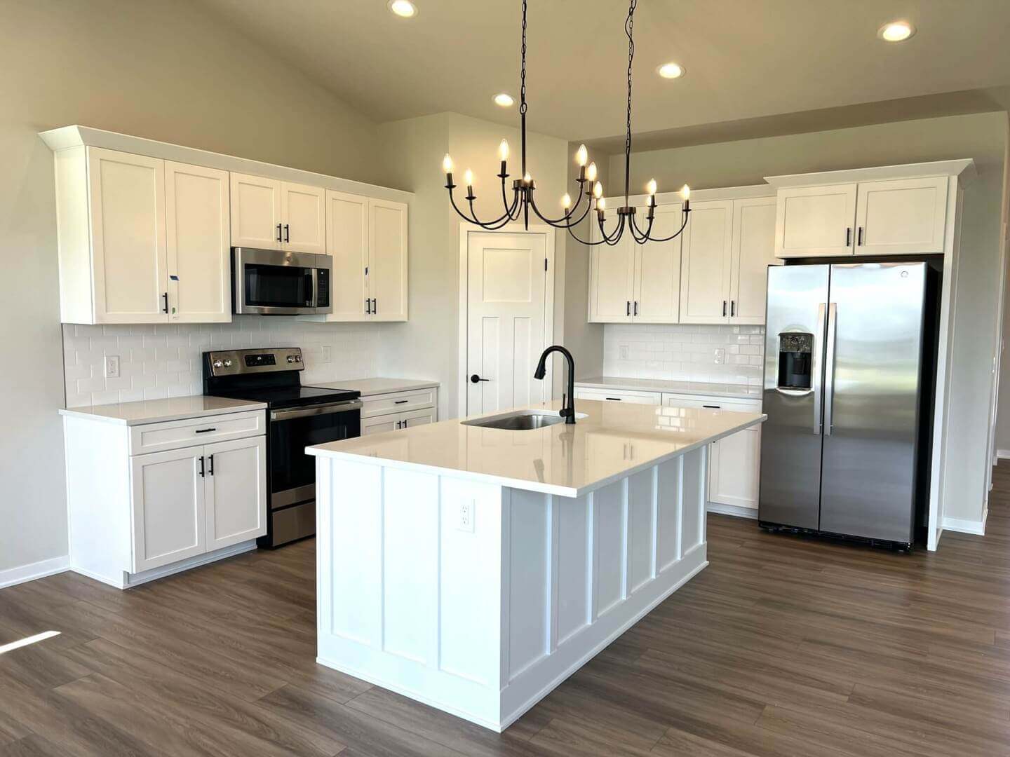 A modern kitchen with white cabinets, stainless steel appliances, and a central island featuring a black faucet and sink. A black chandelier hangs above the island. The floor is light brown wood, and the walls are neutral-colored. Recessed lighting is installed in the ceiling.