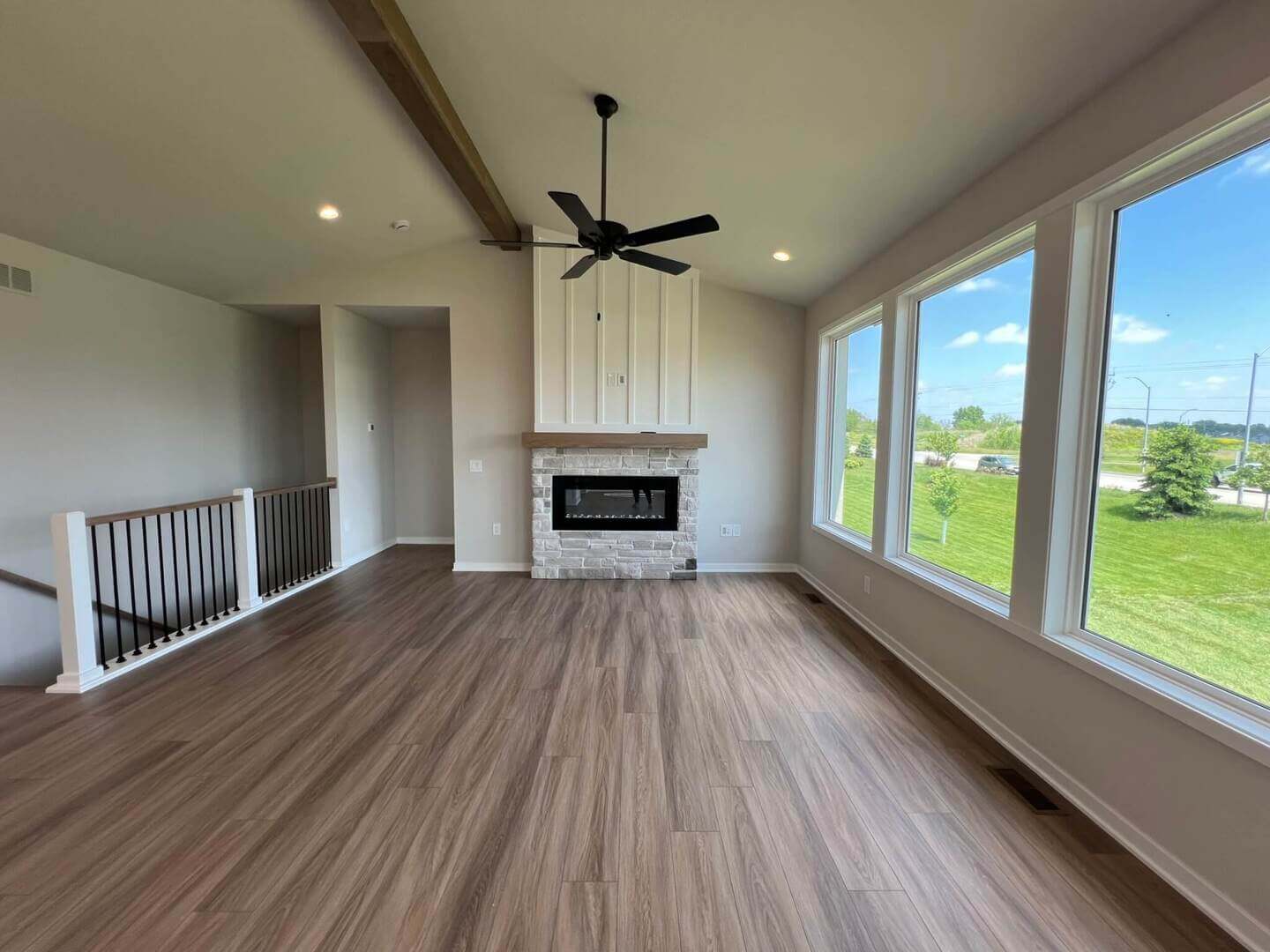 A spacious, unfurnished living room with a high ceiling and large windows offering a scenic outdoor view. The room has hardwood flooring, a white brick fireplace along one wall, a ceiling fan, and a railing separating an adjacent area. Natural light fills the space.