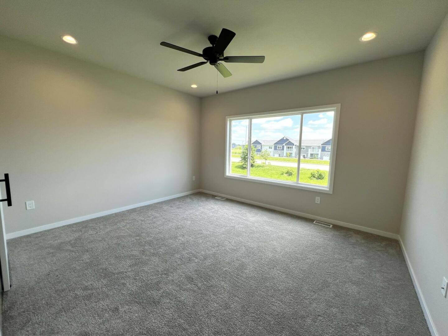 A clean, empty bedroom with light gray walls and carpet. A large window overlooks houses and a grassy field outside. The ceiling features recessed lighting and a black ceiling fan. A dark door handle is visible on the left side of the wall.