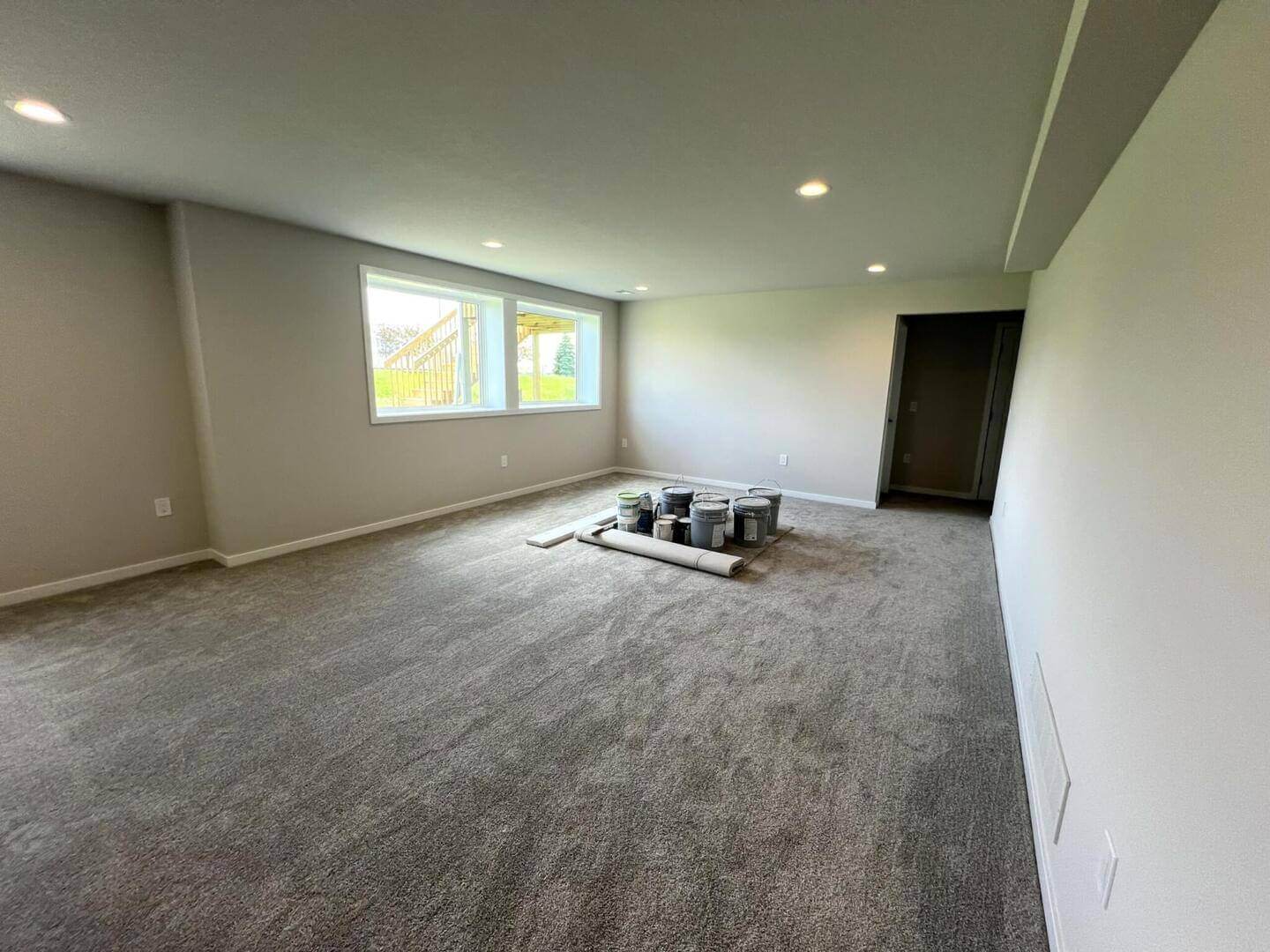 A spacious room with light gray carpet and white walls. Several paint cans and a rolled-up carpet are placed in the center of the floor. A pair of large windows are on the far wall, and a hallway entrance is visible to the right. Recessed ceiling lights are on.