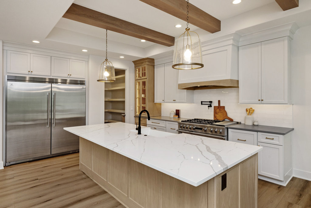 Modern kitchen interior with a white marble island featuring a black faucet. Stainless steel double-door refrigerator and gray cabinetry with brass handles line the walls. Wooden ceiling beams and pendant lights add a rustic touch. Light wood flooring throughout.