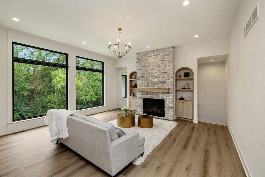 A modern living room with a light gray sofa facing a stone fireplace. The room features large windows with a view of greenery, light wood flooring, and a white rug. Two round gold coffee tables are placed in front of the sofa. Shelves are built into the wall next to the fireplace.