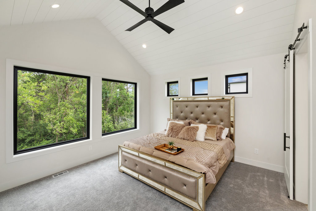 A bedroom with a slanted white paneled ceiling and recessed lighting features a large bed with a beige tufted headboard. Three large windows provide a view of greenery outside. The room has gray carpeted flooring, and a black ceiling fan hangs above the bed.