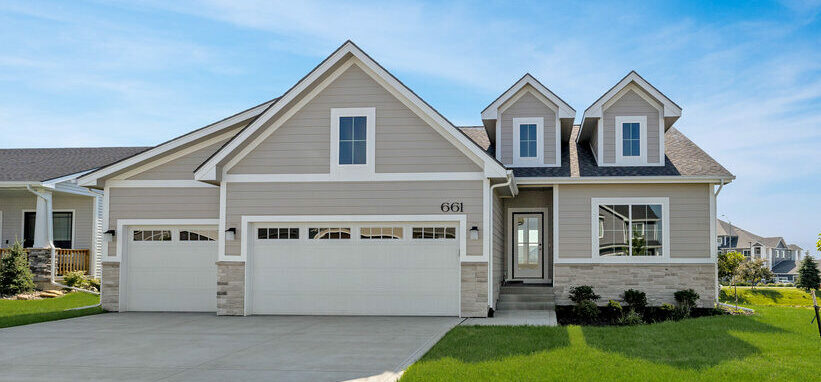 A modern two-story house with beige siding, a three-car garage, and a well-manicured lawn. The home features two dormer windows and is set against a clear blue sky in a suburban neighborhood. A young tree is planted near the driveway on the right side.