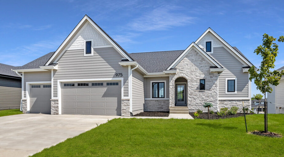 A modern single-story house with a gray exterior, stone accents, and a neatly manicured lawn. The home features a three-car garage with light gray doors and a concrete driveway. A small tree and several small shrubs decorate the front yard. Clear blue sky above.