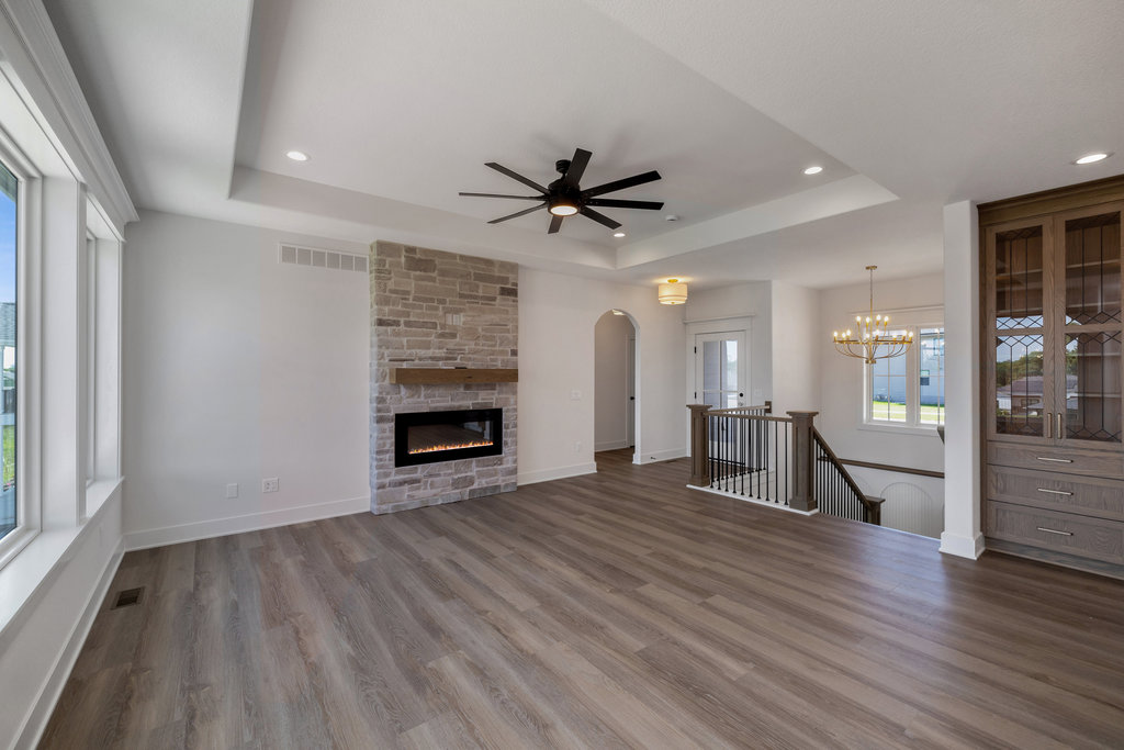 A spacious and modern living room with light-colored walls and wood flooring. It features a stone fireplace, a large ceiling fan, recessed lighting, and a built-in wooden cabinet. There is a staircase with a dark railing leading to the lower level, and a chandelier above.