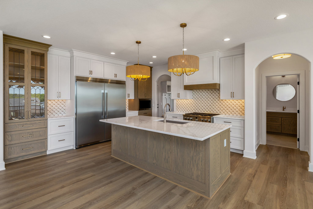 A modern kitchen features a large island with a marble countertop and wooden base, two pendant lights above, white cabinetry, stainless steel appliances including a double-door fridge, and a wood pattern backsplash. The floor is hardwood, with a visible hallway on the right.