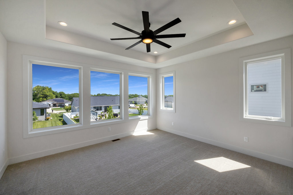 A spacious, empty room with light-colored walls and carpet, featuring large windows offering a view of a suburban neighborhood with houses and greenery. A modern, black ceiling fan is centered on the ceiling above. Natural light illuminates the room.