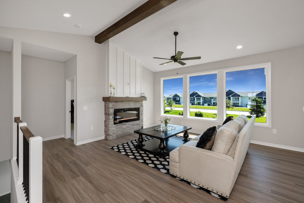 Modern living room with a beige sectional sofa, black and white rug, and a wooden coffee table facing a white shiplap fireplace with a wooden mantel. Large windows offer a view of a bright, suburban neighborhood with houses and greenery. Ceiling fan overhead.