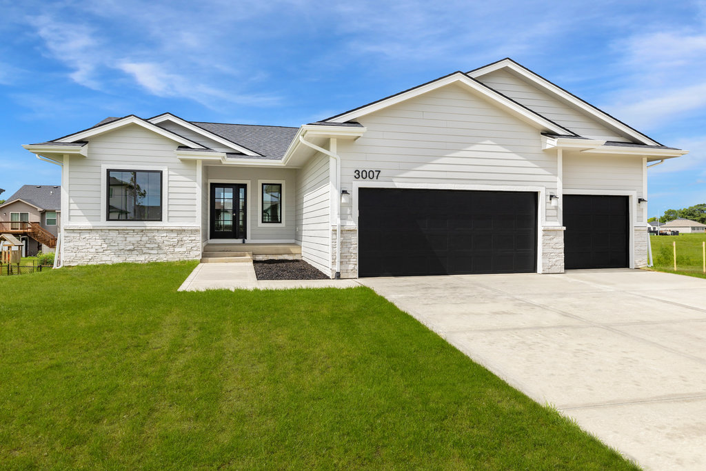 A modern single-story house with white siding, a dark roof, and black garage doors. It has a large driveway and a well-maintained lawn. The sky is clear and blue.