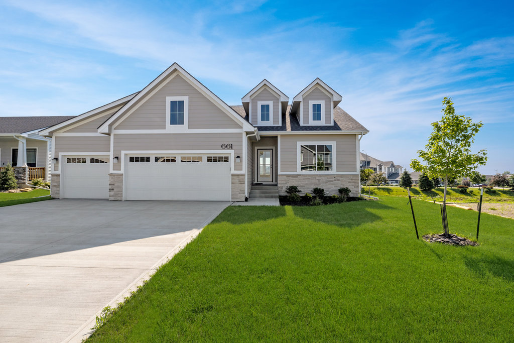 A modern suburban house with a gray exterior, a three-car garage, and dormer windows. It has a well-manicured lawn, a concrete driveway, and a young tree in the front yard under a clear blue sky.