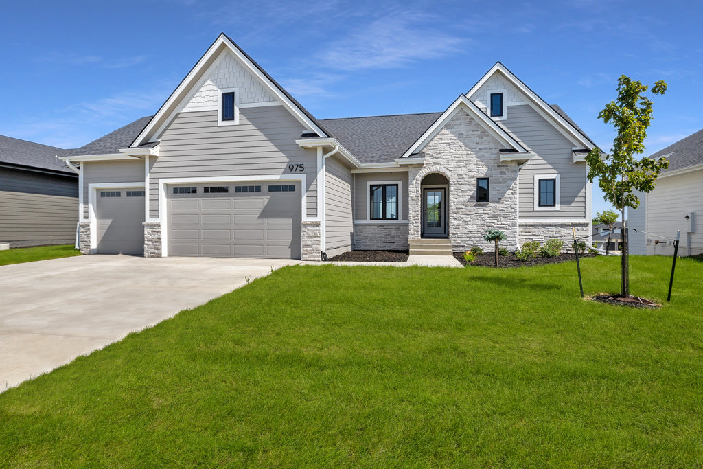 A modern single-story house with a light gray exterior, a dark gray roof, and a stone-accented facade. It features a three-car garage. The yard is well-maintained with lush green grass, a small tree, and minimal landscaping. The sky is clear with no clouds.