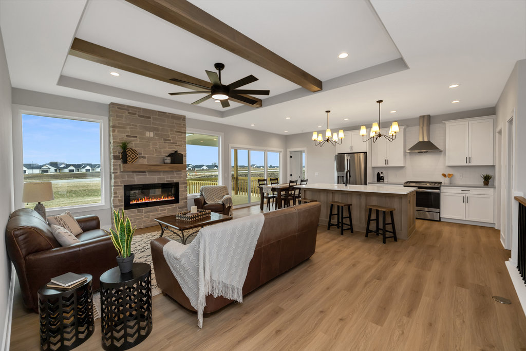 A modern open-concept living room and kitchen area with light wooden floors, a ceiling fan, and recessed lighting. The space features a fireplace, a brown leather sofa, a kitchen island with barstools, white cabinetry, and large windows with a view of a grassy field.