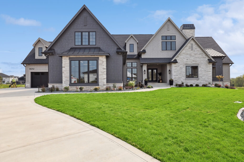 A large, modern two-story house with a mix of gray siding and stone exterior. It features multiple gables, large windows, and a welcoming front porch. The driveway curves in front of the well-manicured lawn under a clear blue sky.