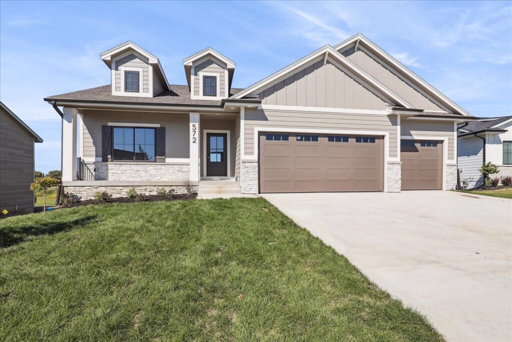 A modern, single-story house with beige siding and stone accents features a two-car garage. The front is landscaped with a small patch of grass and shrubs. Clear blue sky is in the background. House number 572 is displayed by the entrance.