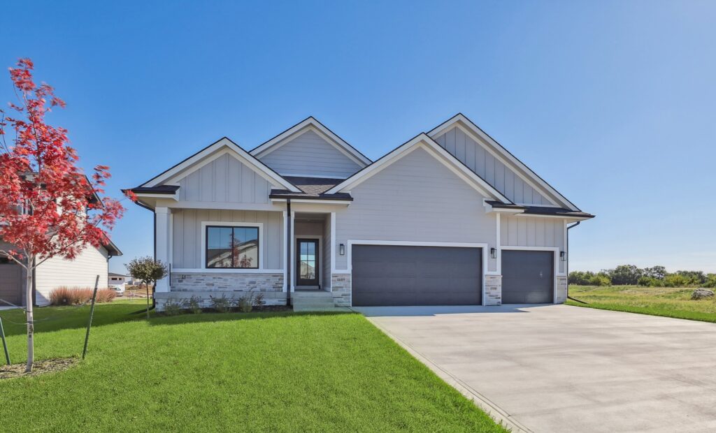 A modern suburban house with a sleek design, featuring a three-car garage, light gray siding, and a well-maintained lawn. A small red tree stands in the front yard against a clear blue sky.