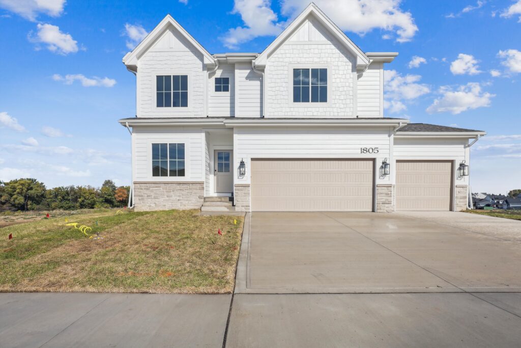 A two-story, modern white house with a gable roof and attached three-car garage. The house features large windows and light gray accents. The driveway is expansive and the yard is newly landscaped, set against a backdrop of blue sky and clouds.