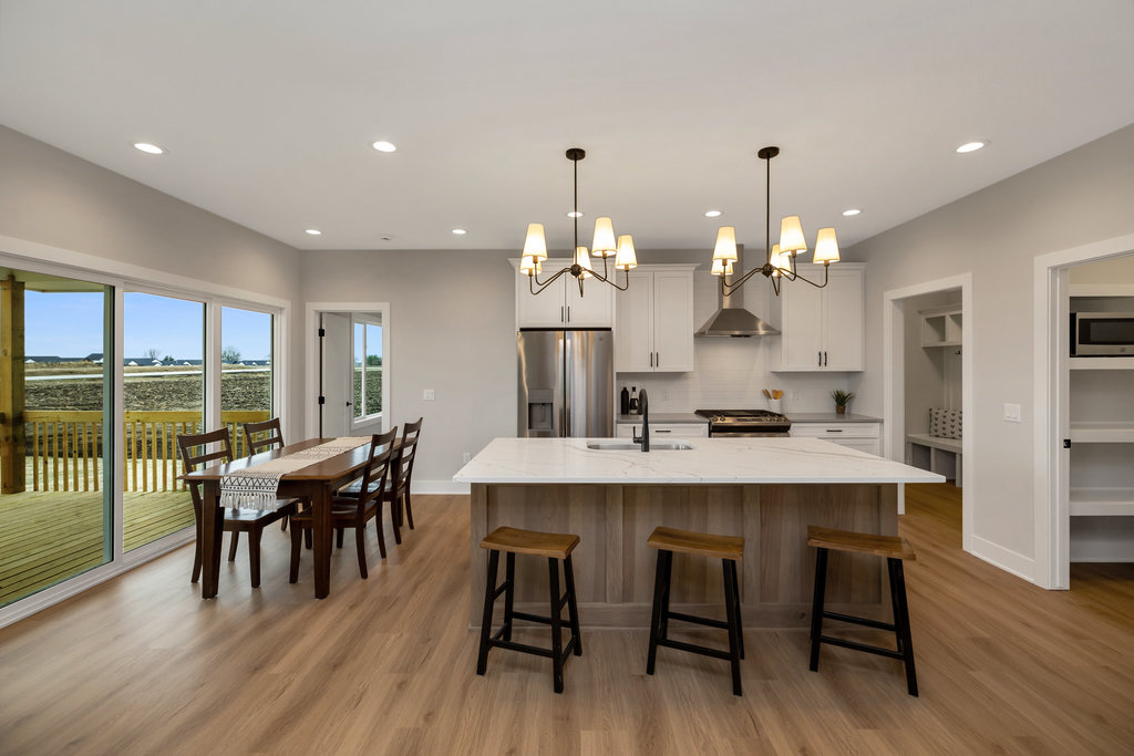 Spacious modern kitchen with a large island featuring three wooden stools. A dining table with chairs is positioned by sliding glass doors. White cabinets, stainless steel appliances, and multiple chandeliers create a bright and welcoming atmosphere.