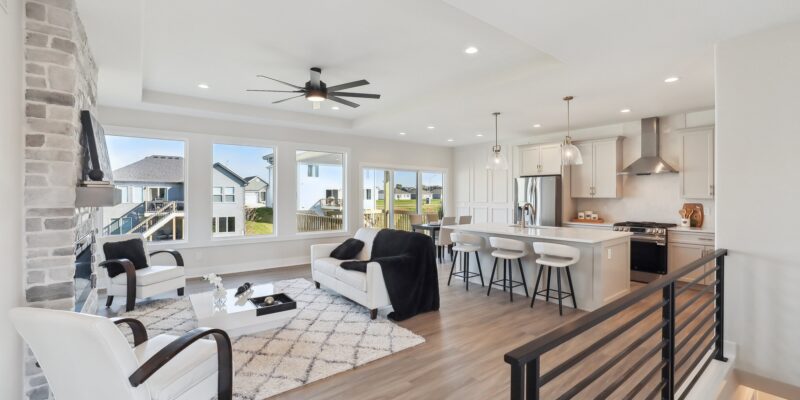 Modern open-concept living room and kitchen with large windows. Features white walls, a stone fireplace, contemporary furniture, a central ceiling fan, and a cozy rug. The kitchen has a breakfast bar with stools and stainless steel appliances.