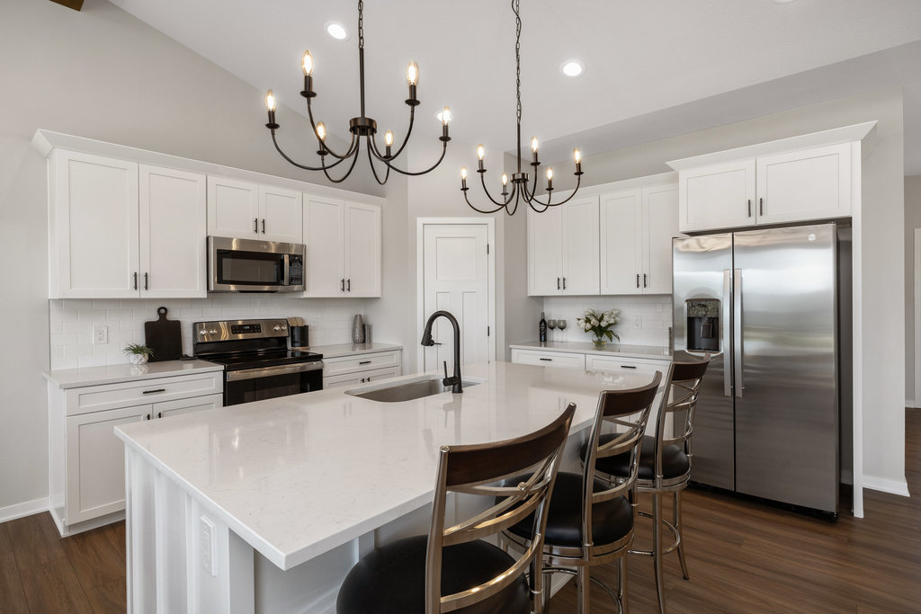 A modern kitchen with white cabinets, a large island, and dark wood chairs. Stainless steel appliances, including a refrigerator and oven, are present. Two black chandeliers hang from the ceiling, and the floor is wooden.