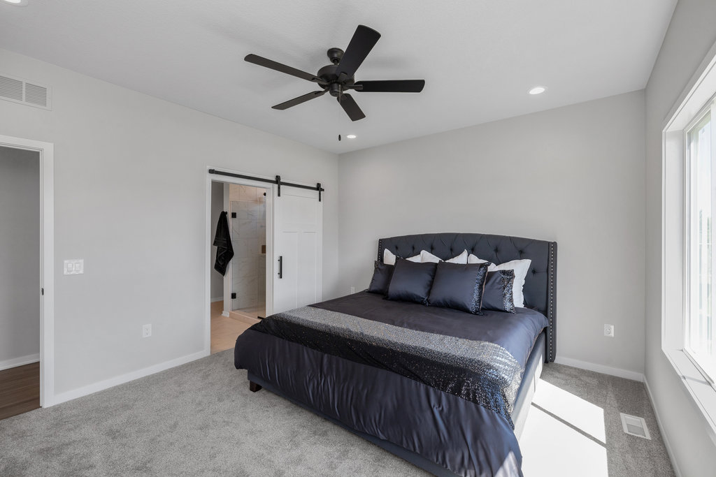 A modern bedroom featuring a large bed with dark bedding and multiple pillows. A ceiling fan is centered above. A sliding barn door reveals a glimpse of a bathroom. The room is lit by natural light from a large window on the right.