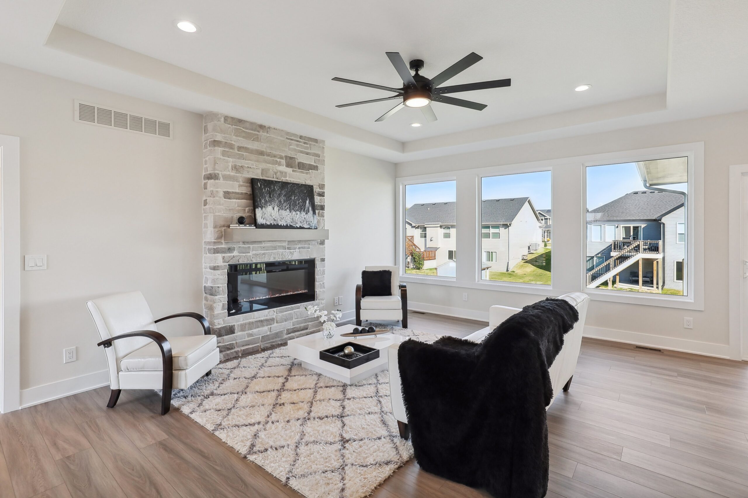 Modern living room with a cozy fireplace, beige walls, and a light wood floor. White armchairs and a black throw blanket are arranged around a small white coffee table on a patterned rug. Large windows reveal a sunny suburban view.