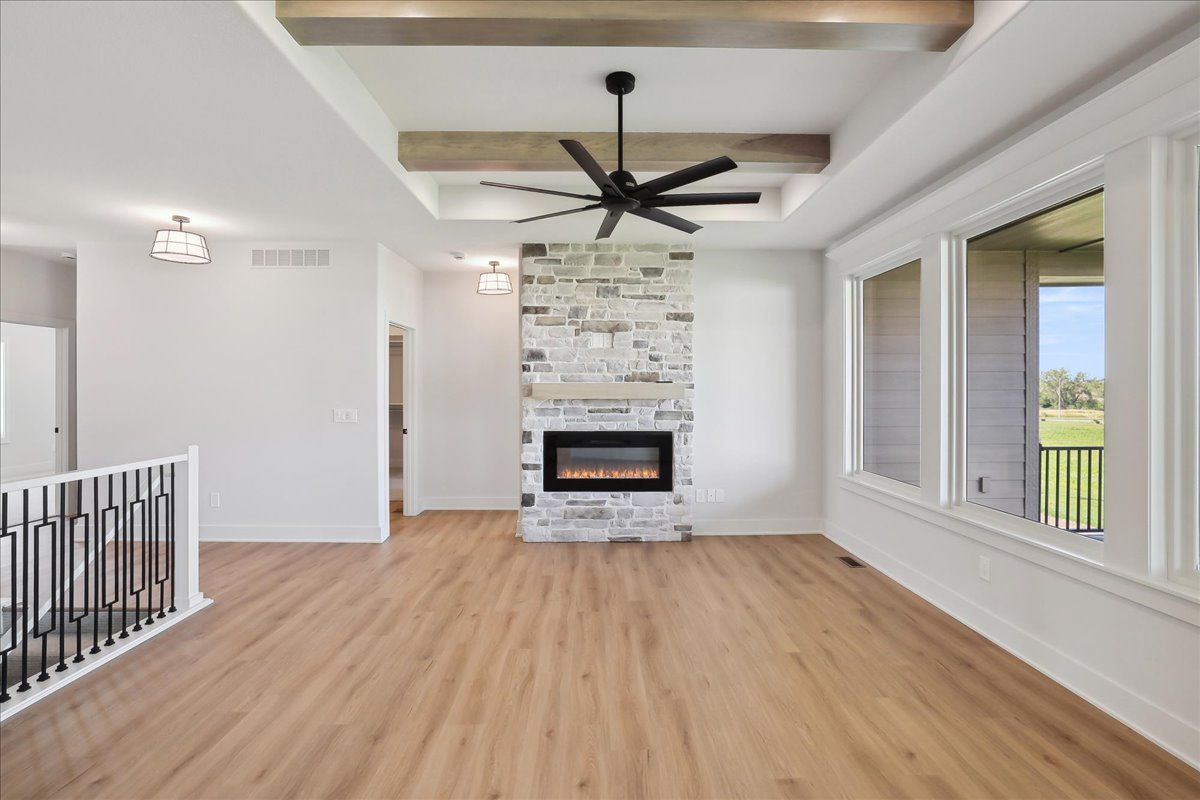 Modern living room with light wood flooring, a stone fireplace, and large windows. The ceiling features wooden beams and a black ceiling fan. Walls are painted in a soft white, creating a bright and airy atmosphere.