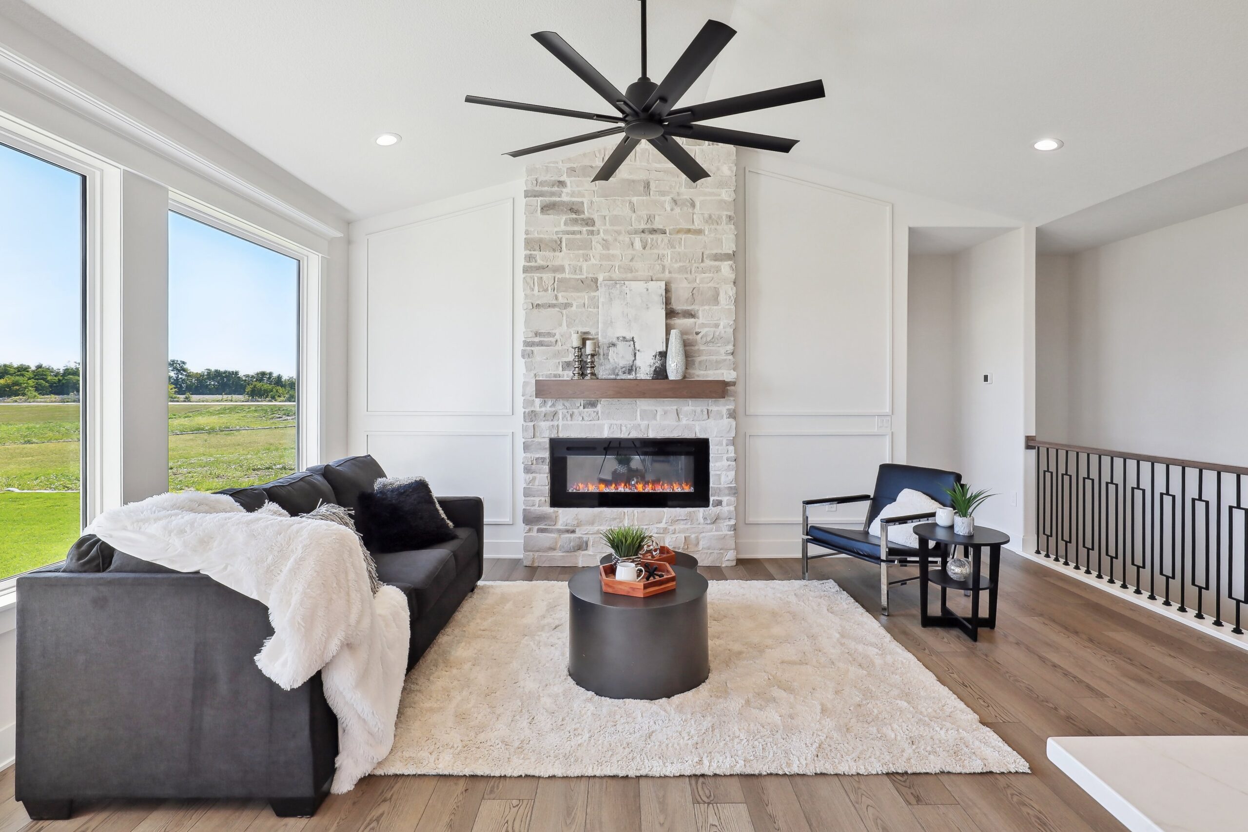 A modern living room with a large window, featuring a black sofa, white fluffy throw, and a stone fireplace with a wooden mantel. There's a round coffee table, two chairs, and a large black ceiling fan. The flooring is light wood.