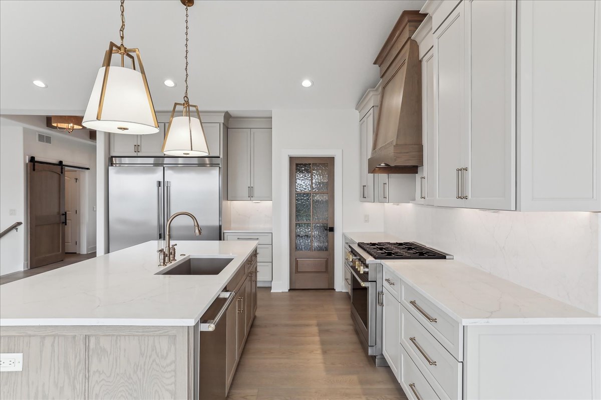 A modern kitchen with light-colored cabinetry, a large island with a sink, and three pendant lights hanging above. The kitchen features a stainless steel refrigerator, a professional-grade oven range with a wooden hood, and a glass-paneled pantry door.