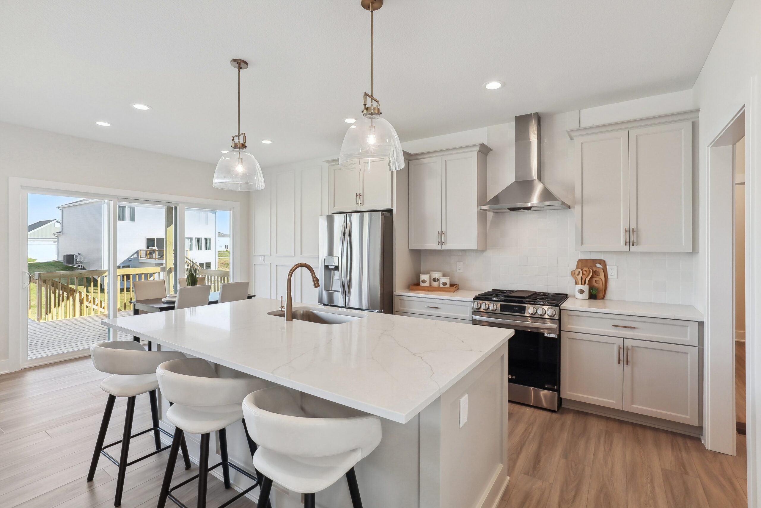 A modern kitchen with a large marble island, white cabinets, stainless steel appliances, and pendant lights hanging above the island. Bar stools line the island, and a sliding glass door leads to an outdoor deck.