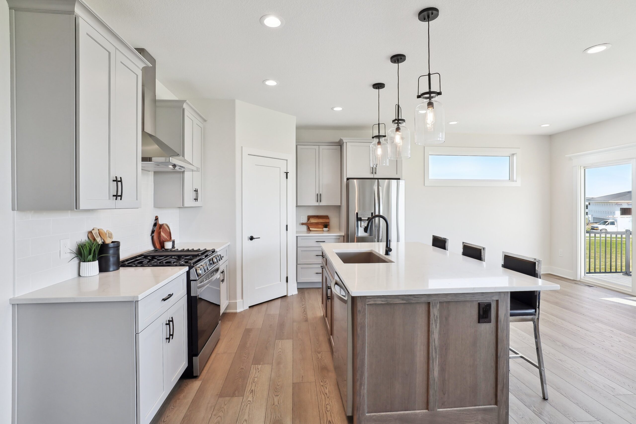 Modern kitchen with stainless steel appliances, white cabinets, and a large island featuring a wooden base and white countertop. Three pendant lights hang above the island, which is accompanied by bar stools. Light pours in from a large window.