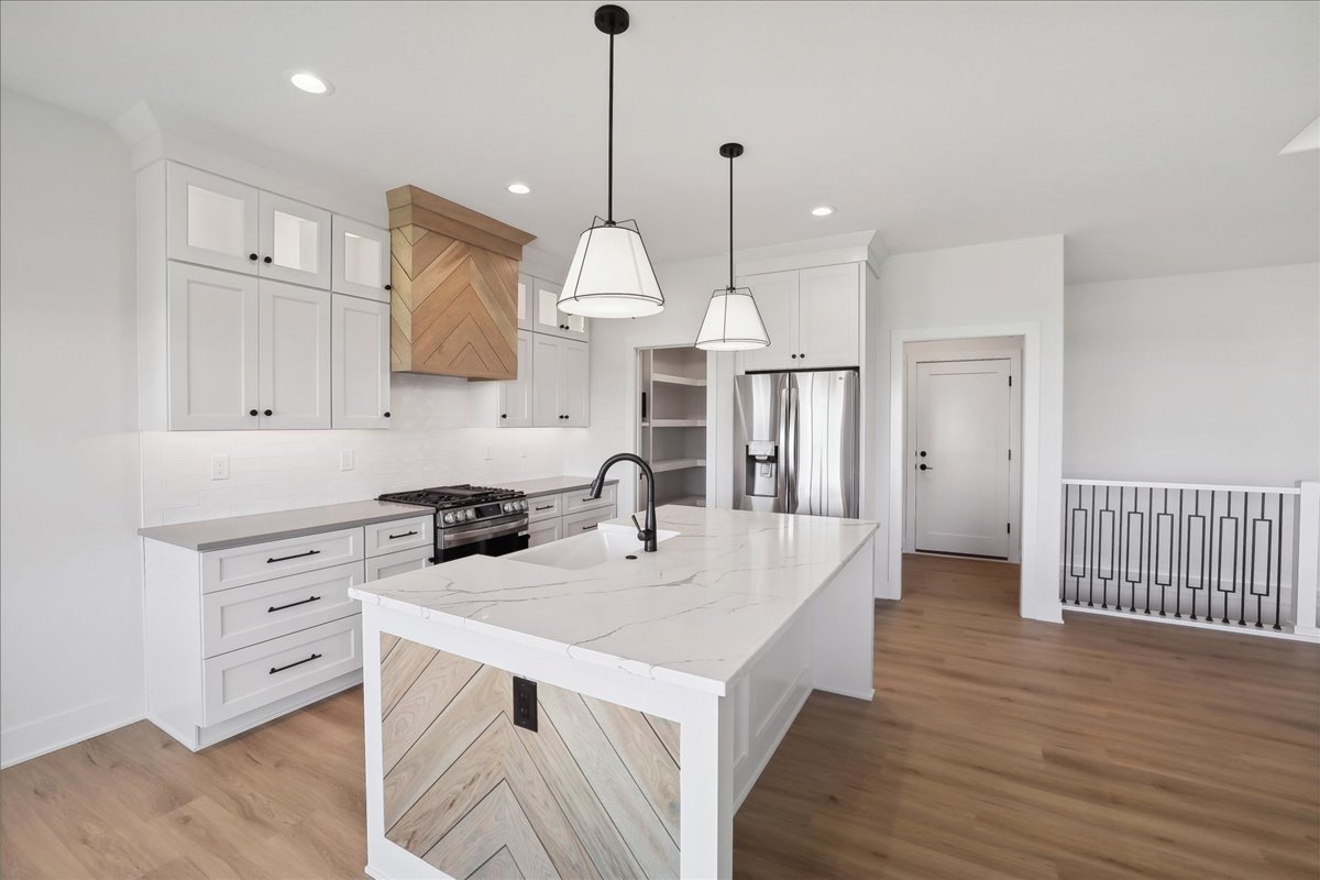 Modern kitchen with white cabinets, a large marble island, and stainless steel appliances. The island features chevron wood paneling. Pendant lights hang above the island, and the space has light wood flooring and a farmhouse-style hood.