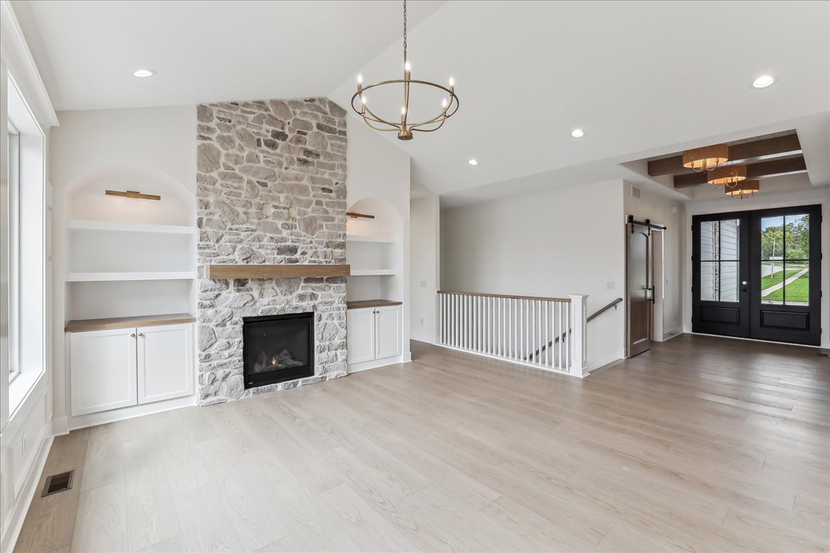 A spacious, modern living room with a stone fireplace flanked by built-in shelves, light wood floors, and a chandelier. In the background, there's an entryway with double doors and a view of a staircase.