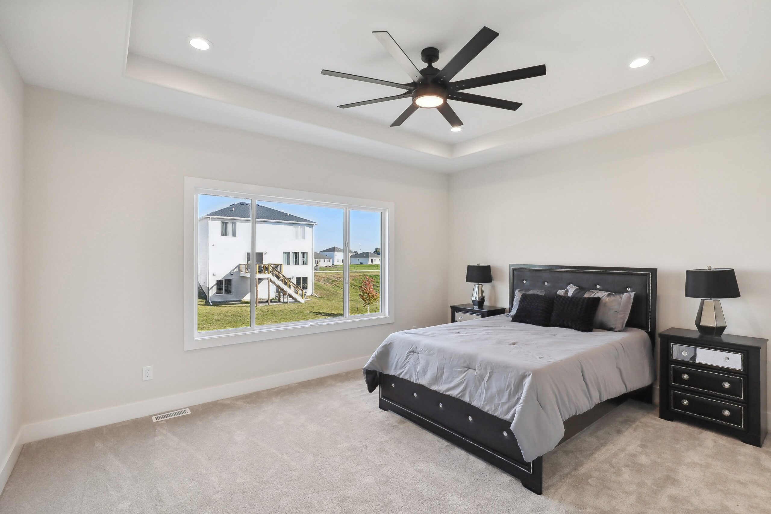 A modern, neutral-toned bedroom with a large window showing an outdoor view. The room features a bed with gray bedding, two black nightstands with lamps, a ceiling fan, and carpeted flooring.