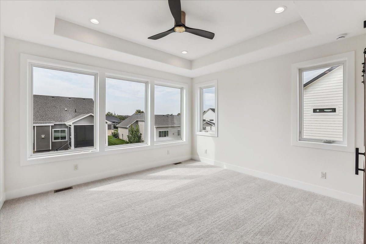 A bright, empty room with beige carpet and large windows on two walls showing a suburban neighborhood. A black ceiling fan is centered in the white ceiling, which features recessed lighting. Walls are painted white, creating a clean and airy feel.