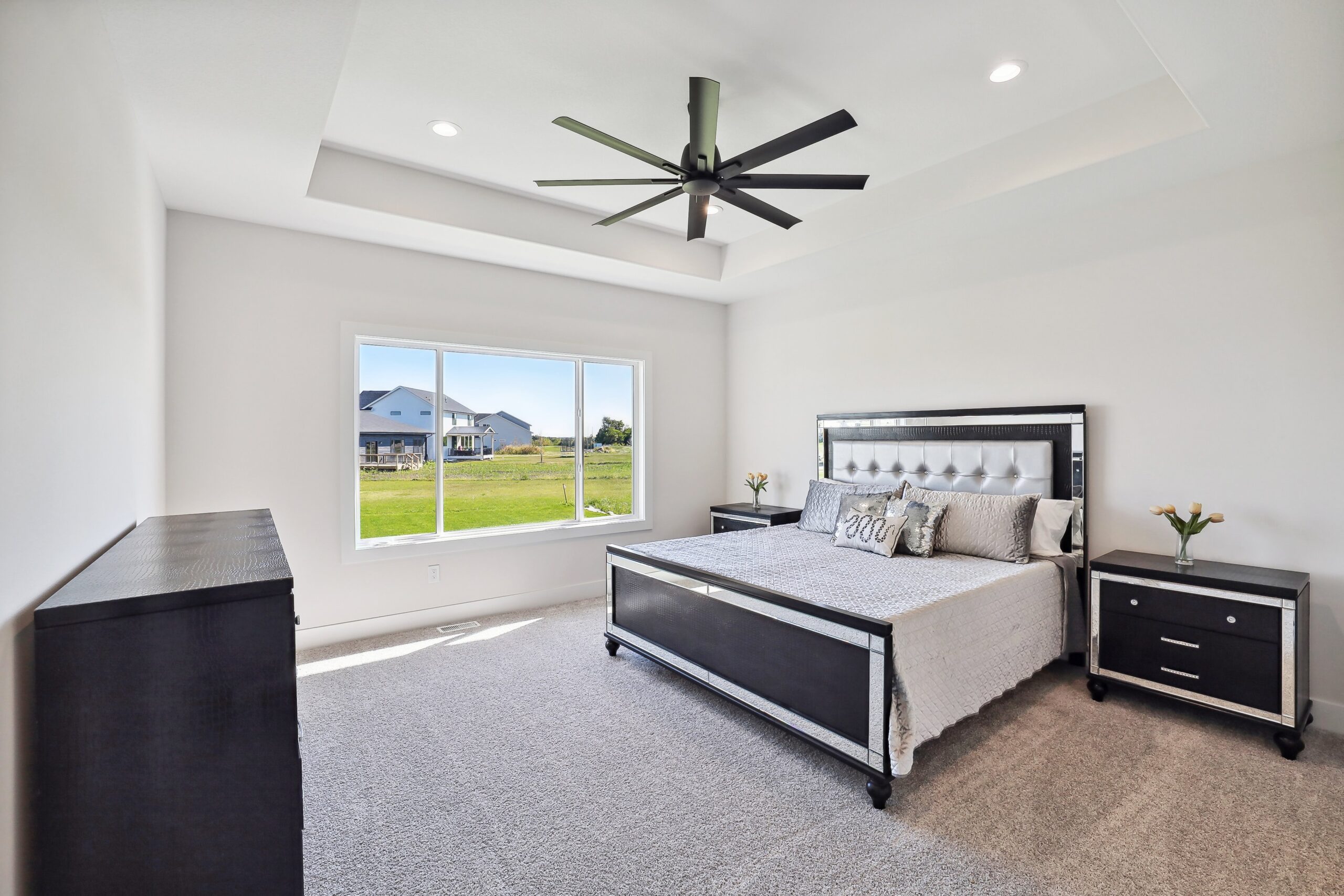 A modern bedroom with a large bed featuring black and white bedding. A ceiling fan hangs from a recessed ceiling. There's a window with a view of grassy fields and a house in the distance. The room has a dresser and two matching nightstands.